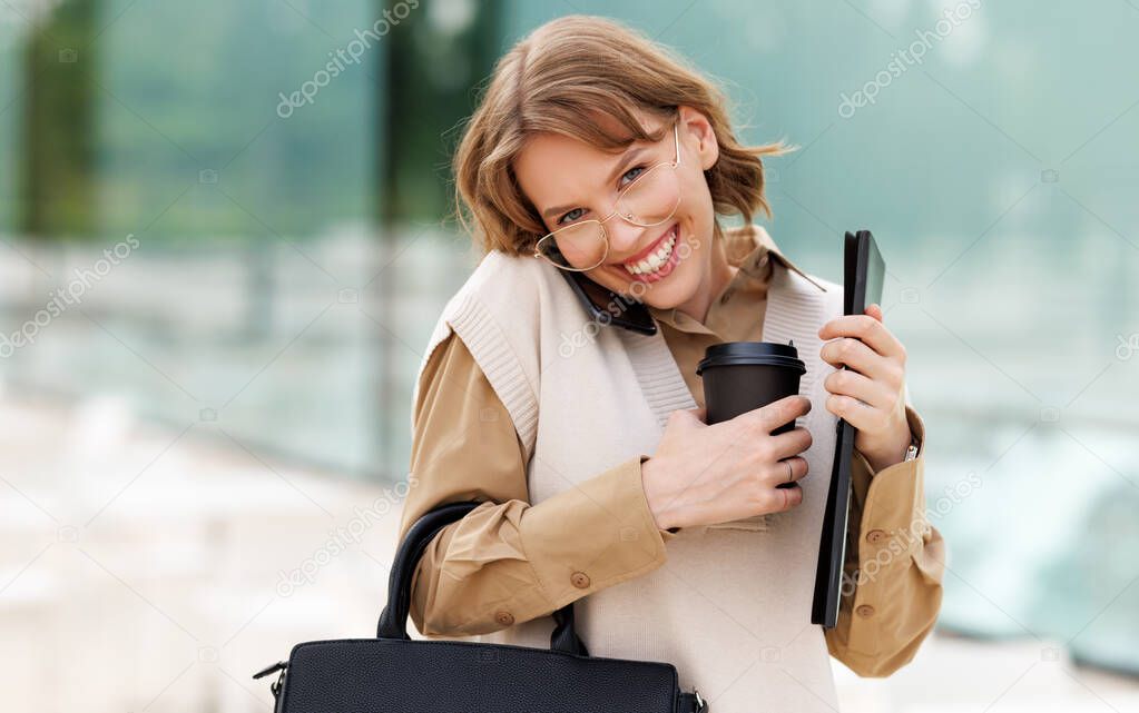 Young smiling female stands outside in city park with coffee while speaking on mobile phone