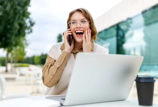 Ofis çalışanı kahve ve dizüstü bilgisayarla terasta oturmuş gülümseyen cep telefonuyla konuşuyor. — Stok fotoğraf