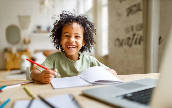 Hymyilevä afrikkalainen amerikkalainen lapsi koulupoika opiskelee verkossa kannettavan kotona — kuvapankkivalokuva