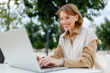 Gözlüklü güzel kız terasta dizüstü bilgisayarla oturuyor.