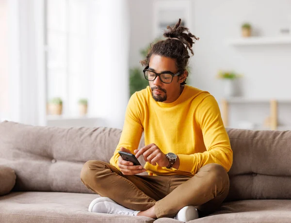 Jonge gefocuste serieuze Afrikaanse Amerikaanse man ontspannen thuis met smartphone, surfen internet — Stockfoto