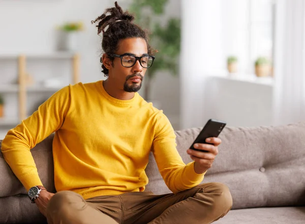 Jonge gefocuste serieuze Afrikaanse Amerikaanse man ontspannen thuis met smartphone, surfen internet — Stockfoto