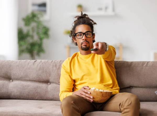 Sıkılmış Afrikalı Amerikalı adam evde dinlenirken uzaktan kumandayla TV 'yi değiştiriyor. — Stok fotoğraf