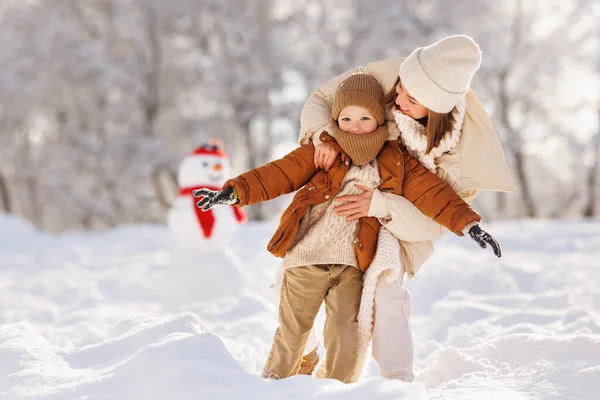 . счастливая семья мать и сын играют, обнимаются и веселятся рядом со снеговиком на зимней прогулке в парке — стоковое фото
