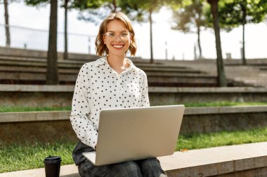 Resmi kıyafetli neşeli genç bir kadın dışarıda bankta otururken bilgisayarla uzaktan çalışıyor.