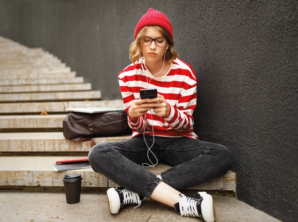 Moody chateado estudante menina em fones de ouvido sentado fora e mensagens de texto amigos no telefone celular — Fotografia de Stock