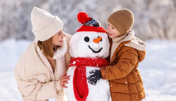 Alegre joven madre haciendo muñeco de nieve con hijo mientras juegan juntos en el parque de invierno nevado — Foto de Stock