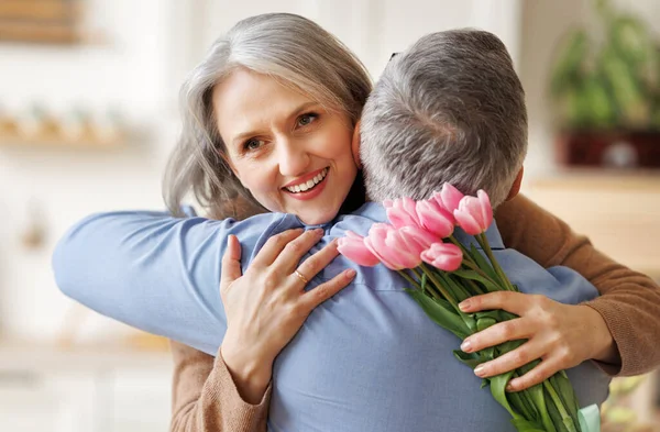 Feliz mujer mayor que recibe el ramo de flores de primavera tulipanes de marido en el Día de la Mujer en casa —  Fotos de Stock