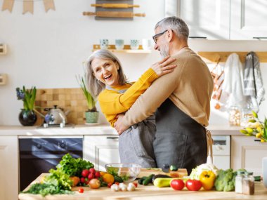 Mutlu yaşlı çift karı koca, mutfakta birlikte yemek pişirirken kucaklaşıp dans ediyor.