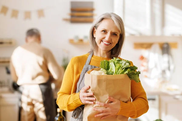 幸せなお年寄りの女性は台所で野菜の袋を手に — ストック写真