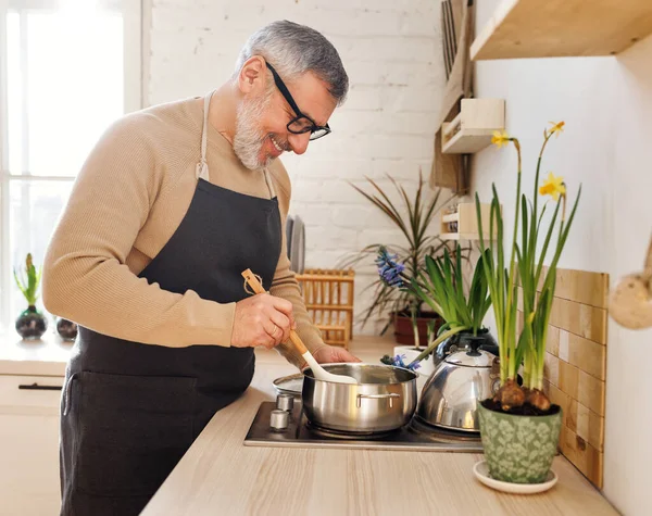 Счастливый пожилой человек в фартуке готовит дома на кухне, стоит рядом с плитой и перемешивает соус в кастрюле — стоковое фото