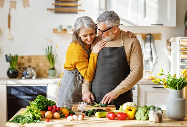 Glückliches älteres Ehepaar in Schürzen kocht gemeinsam gesundes Essen in gemütlicher Küche zu Hause — Stockfoto