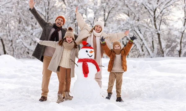 Happy family in warm clothes laughing merrily and raising hands up while making snowman together — Stock Photo, Image