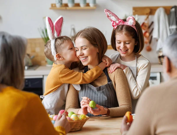 Lindos niños pequeños abrazando y besando a la joven madre feliz mientras pintan huevos de Pascua con la familia — Foto de Stock