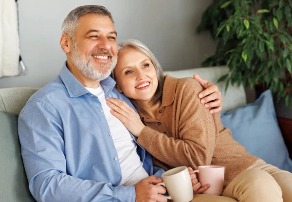 Amar a la pareja de ancianos de la familia bebiendo té y abrazando mientras se relaja en el acogedor sofá en casa —  Fotos de Stock