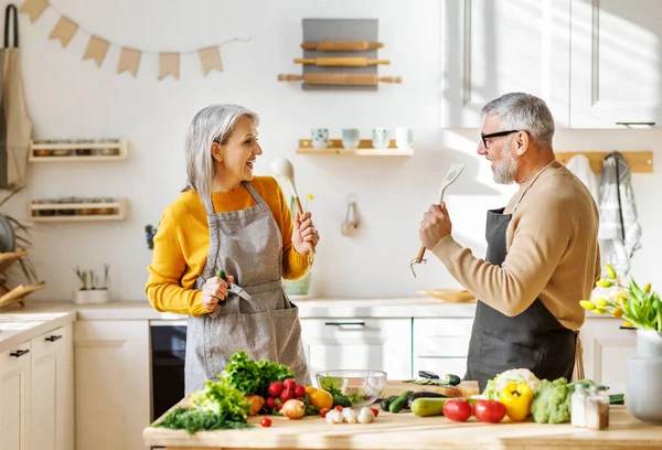 Радостная пожилая пара весело танцует и поет, готовясь вместе на кухне — стоковое фото
