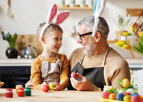 Любящий дедушка и ребенок в повязках на ушах, украшающих яйца на Пасху на светлой уютной кухне — стоковое фото