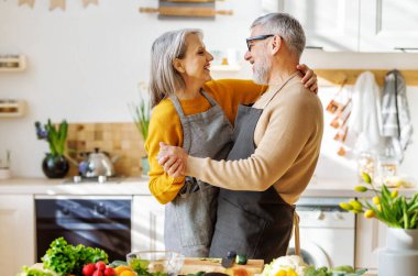 Mutlu yaşlı çift karı koca, mutfakta birlikte yemek pişirirken kucaklaşıp dans ediyor.