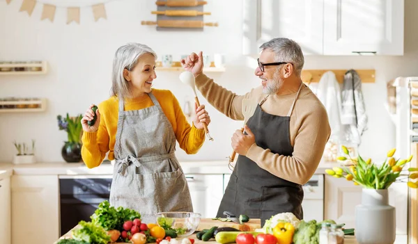Neşeli yaşlı çift mutfakta birlikte yemek pişirirken dans edip şarkı söylerken eğleniyor. — Stok fotoğraf