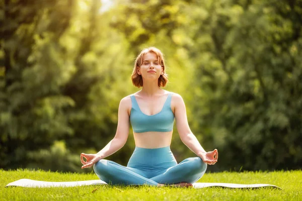 Menina desportiva adorável feliz realizando ioga enquanto sentado no tapete na posição de lótus na natureza — Fotografia de Stock