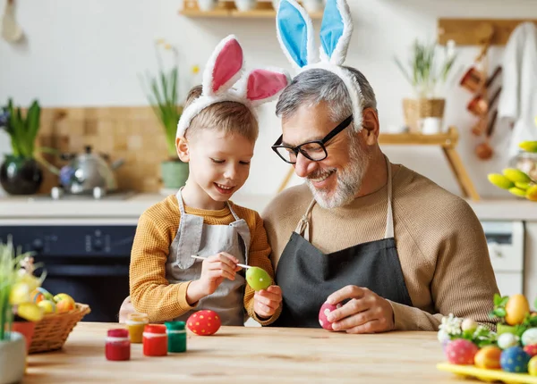 在轻盈舒适的厨房里，可爱的祖父和孩子戴着兔子耳坠，装饰复活节彩蛋 — 图库照片