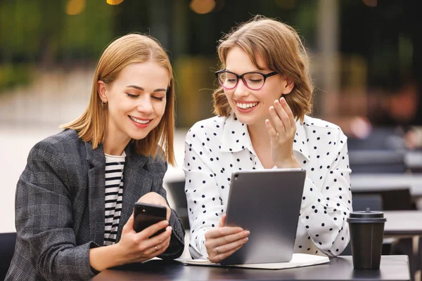 Emocionado exitosas mujeres de negocios en la cafetería al aire libre mirando la tableta digital y el teléfono inteligente con expresión de cara feliz —  Fotos de Stock
