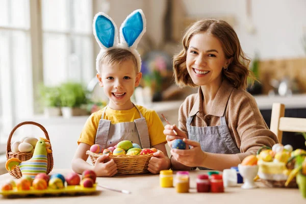 Amar a mãe jovem ensinando o filho pequeno feliz para decorar ovos de Páscoa enquanto sentado na cozinha — Fotografia de Stock