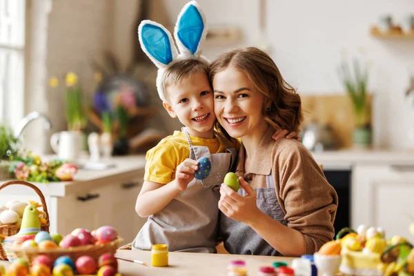 Любящая молодая мать учит счастливого маленького сына украшать пасхальные яйца, сидя на кухне — стоковое фото