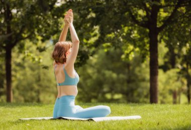 Genç sakin kadın parkta yoga minderinin üzerinde oturuyor. Nilüfer pozisyonunda. Elleri havada ve meditasyon yapıyor.