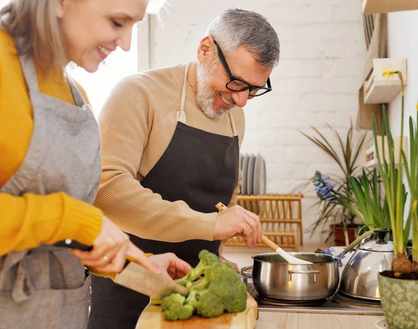 Feliz alegre pareja de ancianos en delantales preparando sopa vegetariana juntos en la acogedora cocina —  Fotos de Stock