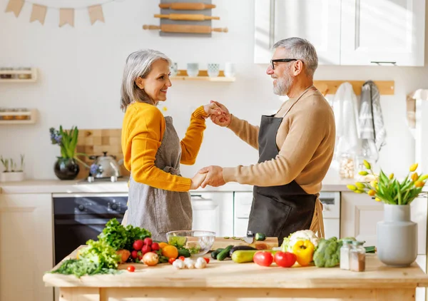 Счастливая пожилая пара муж и жена обнимаются и танцуют, готовясь вместе на кухне — стоковое фото