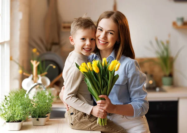 Mujer joven madre con ramo de flores abrazando hijo mientras recibe felicitaciones en el día de las madres — Foto de Stock