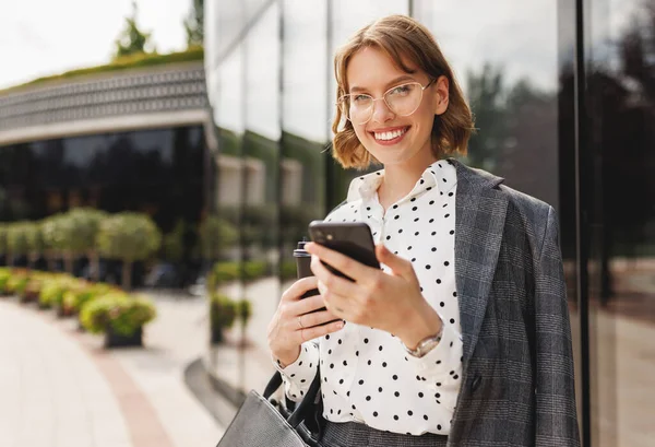 Positiv affärskvinna med ta bort kaffe med mobiltelefon när du står på solig gata — Stockfoto