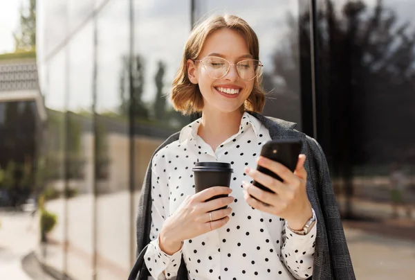 Positiv affärskvinna med ta bort kaffe med mobiltelefon när du står på solig gata — Stockfoto