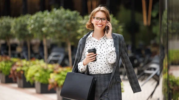 Atractiva dama de negocios haciendo llamada mientras camina por la calle de la ciudad con llevar café en la mano —  Fotos de Stock