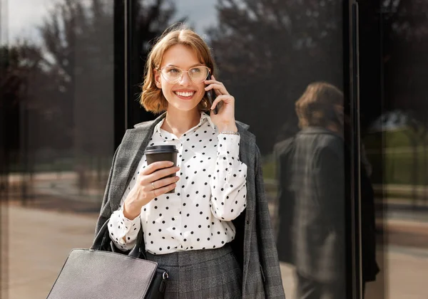 Senhora de negócios atraente fazendo chamada enquanto caminhava ao longo da rua da cidade com tomar café na mão — Fotografia de Stock