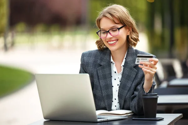 Cheerful business lady with credit bank card paying online while sitting at cafe table with laptop, — Stock Photo, Image