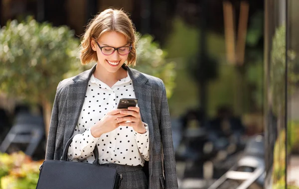 Jonge vrolijke zakenvrouw buiten staan in zonnige dag en kijken naar het scherm van de smartphone — Stockfoto