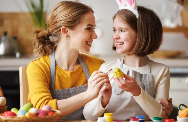 Loving young mother teaching happy kid daughter to decorate Easter eggs while sitting in kitchen — Stock Photo, Image