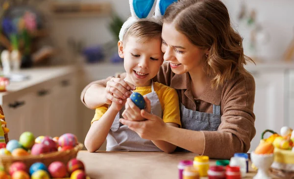 Amar joven madre enseñando feliz niño pequeño hijo para decorar los huevos de Pascua mientras está sentado en la cocina — Foto de Stock