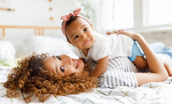 Happy african american mother gently hugs laughing baby daughter in bed — Stock Photo, Image