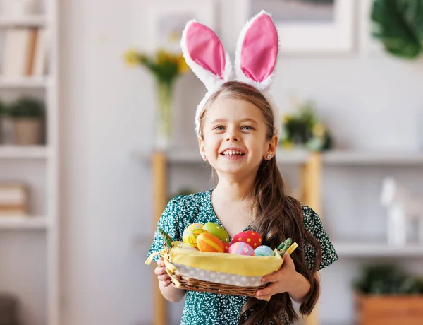 Niña positiva con orejas de conejito sonriendo y sosteniendo la cesta de Pascua en las manos en casa — Foto de Stock