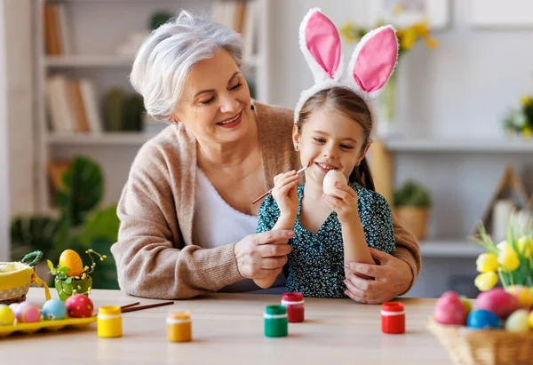 Sevgi dolu büyükanne, küçük mutlu kıza mutfakta otururken paskalya yumurtalarını süslemeyi öğretiyor. — Stok fotoğraf
