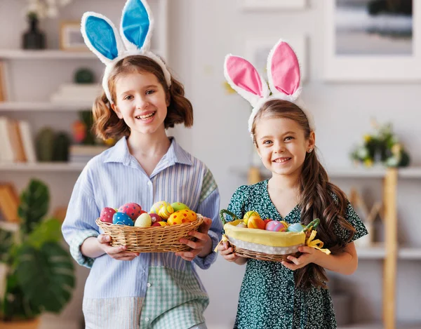 Duas meninas positivas com orelhas de coelho sorrindo e segurando cesta de Páscoa em mãos em casa — Fotografia de Stock