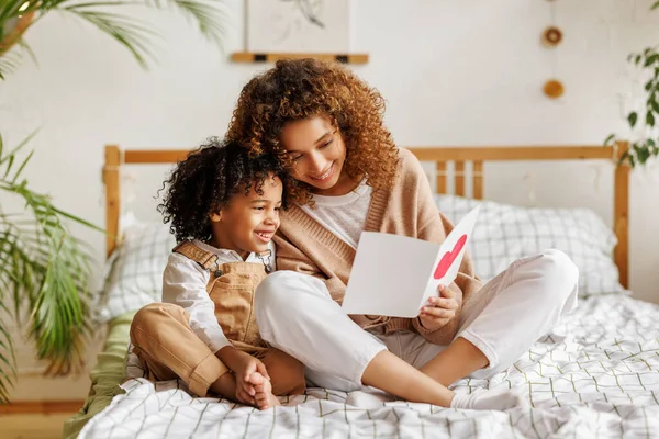 Filho bonito dando a sua mãe cartão postal cumprimentando no dia das mães, comemorando juntos em casa — Fotografia de Stock