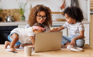 Genç sinirli Afro-Amerikan anne, evden iki küçük çocukla çalışırken kulaklarını kapatıyor ve laptop ekranına bakıyor, gürültücü çocuklar tarafından işten uzaklaştırılıyor.