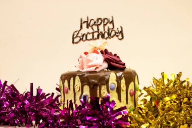 Çikolata parçalarıyla süslenmiş doğum günü pastası ve mutlu yıllar metni, fındık ve güllerle süslenmiş pembe ve altın renkli süsler.