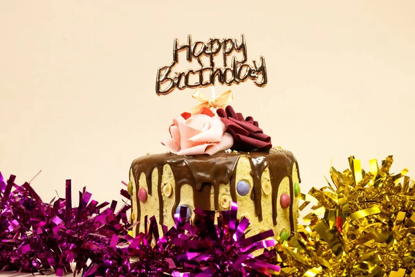 Çikolata parçalarıyla süslenmiş doğum günü pastası ve mutlu yıllar metni, fındık ve güllerle süslenmiş pembe ve altın renkli süsler.