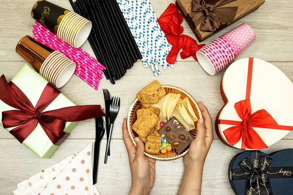 happy birthday concept, hands woman holding Chocolate Candy plate and straw and chips potato, cookies and disposable cups with plastic forks and  gift boxes on wood table