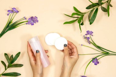 Makyaj cilt bakımını kaldırır. Yüz derisi ya da makyaj malzemesi için plastik bir şişede tonik ve pembe arka planda pamuk tutan eller. Hijyen malzemeleri, güzellik aletleri ve ten, çiçekler.